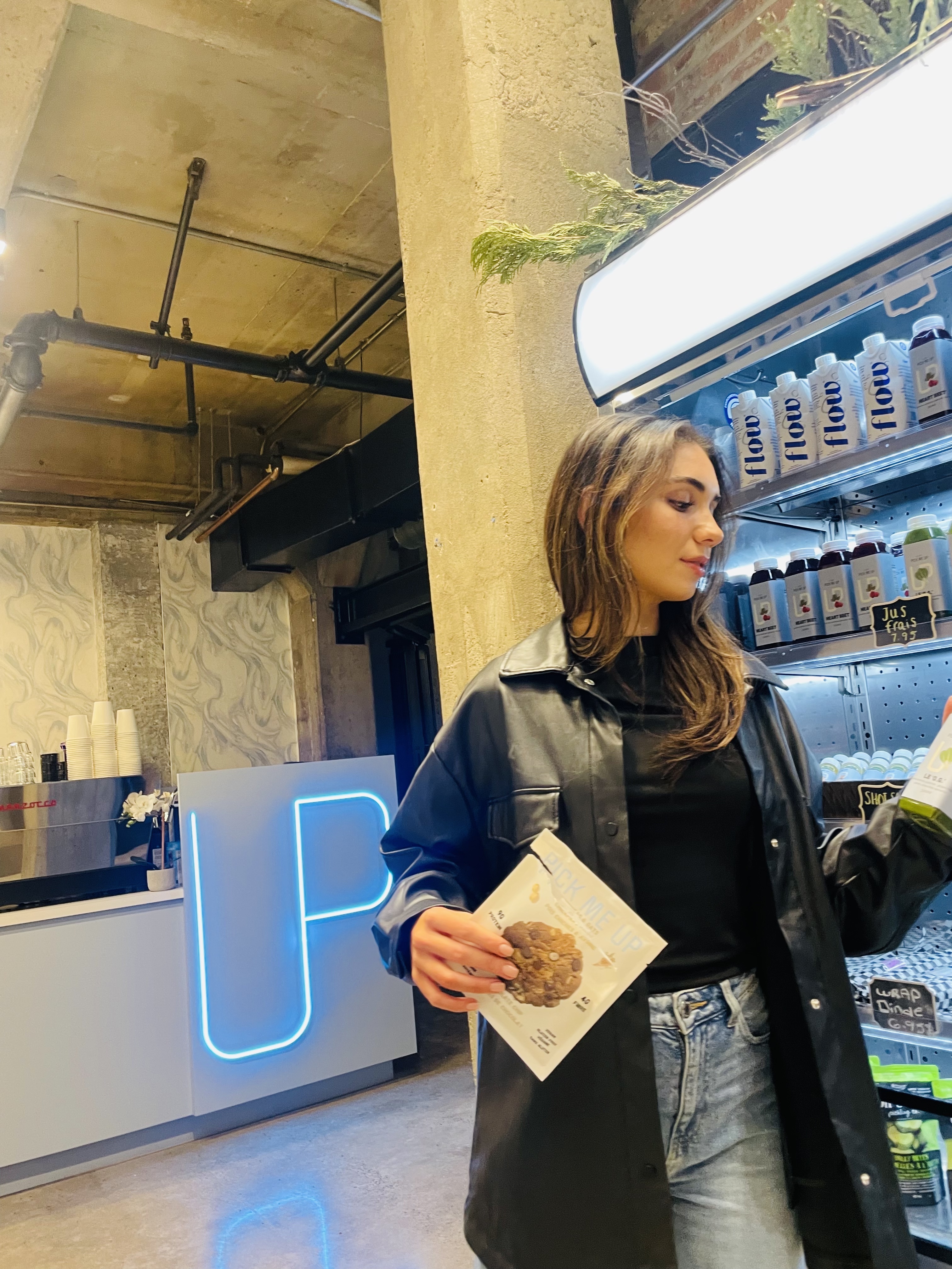 Woman in a black leather jacket holding a Pick Me Up cookie package in a café